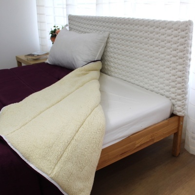 Cama individual com cabeceira acolchoada branca, manta creme felpuda, cobertor roxo e almofada cinza, numa divisão clara com mesa lateral