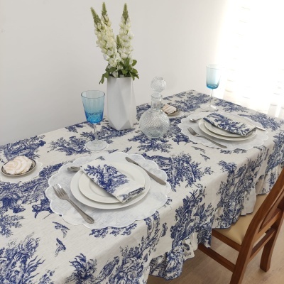 Mesa de madeira posta com toalha e guardanapos de algodão branco com padrão azul, pratos brancos, copos azuis, garrafa de vidro transparente e vaso branco com flores brancas.