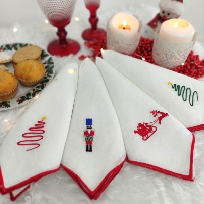 Guardanapos de tecido branco com bordas vermelhas e bordados natalícios em superfície branca, velas acesas e copos vermelhos ao fundo.