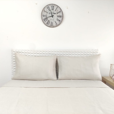 Cama com lençol e almofadas cremes, cabeceira trançada branca e relógio de parede com números romanos.