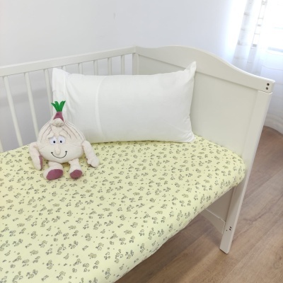 Berço branco com almofada branca, lençol amarelo claro com padrão floral cinza e boneco de pelúcia em forma de alho