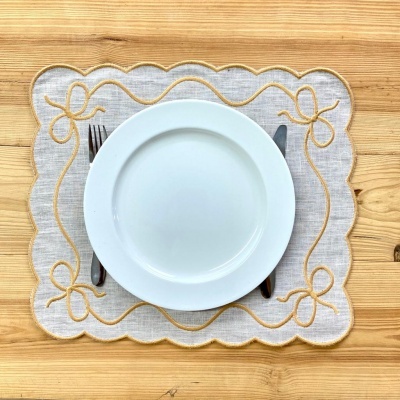 Jogo americano cinza com bordado amarelo, prato branco e talheres numa mesa de madeira.