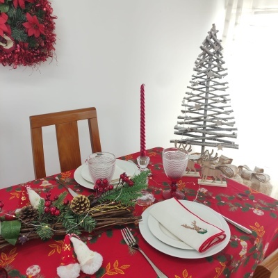 Mesa de jantar decorada para Natal com toalha vermelha, centro de mesa com pinhas e frutos vermelhos, velas vermelhas, louça branca, e mini árvore de Natal de madeira