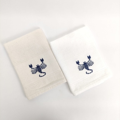 Duas toalhas bege e branca com bordado azul de escorpião sobre fundo branco
