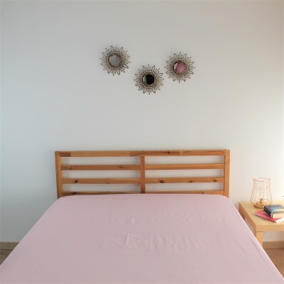 Cama com cabeceira de madeira clara e colchão rosa, mesa de cabeceira e espelhos decorativos na parede branca