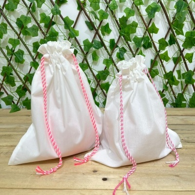 Bolsas de algodão brancas com cordões rosa e branco sobre mesa de madeira clara com fundo de folhas verdes