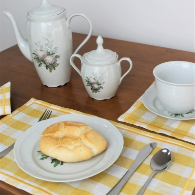 Conjunto de loiça branca com padrão floral e pão numa mesa com toalhas amarelas xadrez