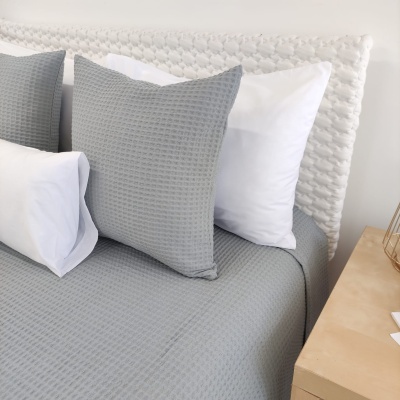 Cama com capa de edredão cinzenta em padrão em relevo e almofadas brancas e cinzentas junto a cabeceira branca trançada