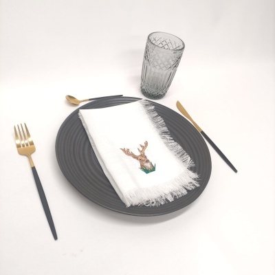 Mesa posta com prato cinzento, guardanapo branco com bordado de veado, copo cinzento e talheres pretos e dourados