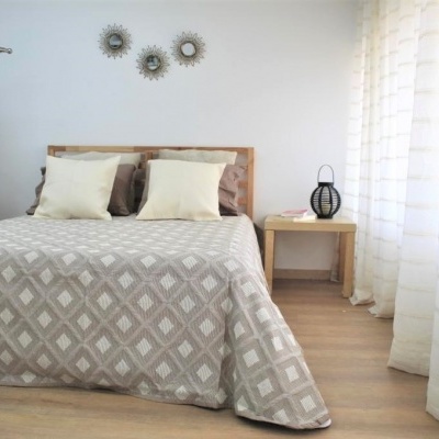 Quarto com cama de madeira, colcha de padrão geométrico branco e cinza, almofadas bege e mesa de cabeceira com abajur preto