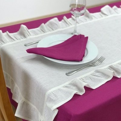 Mesa posta com toalha branca com folhos sobre toalha magenta, prato branco com guardanapo magenta, talheres e copo de vinho