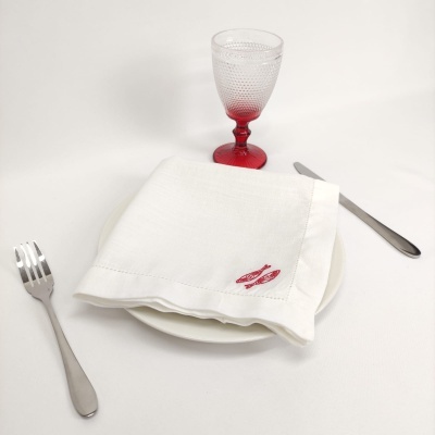 Mesa posta com guardanapo branco bordado, talheres prateados, prato branco e copo de vidro vermelho