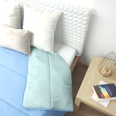 Cobertor azul claro e muito claro sobre cama branca com cabeceira branca em trama, almofadas brancas e bege, mesa de cabeceira de madeira clara com suporte metálico dourado e livro.