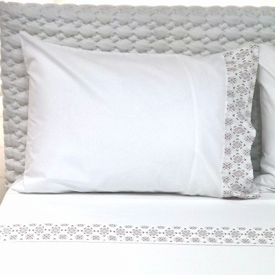 Conjunto de cama branco com detalhes bordados geométricos e cabeceira acolchoada cinza.