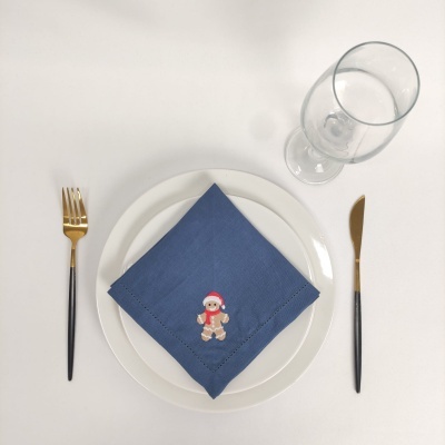 Conjunto de jantar com prato branco, guardanapo azul bordado com boneco de gengibre, talheres dourados e pretos, copo de vidro