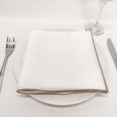 Guardanapo branco com borda castanha sobre prato branco numa mesa posta com talheres e copo.