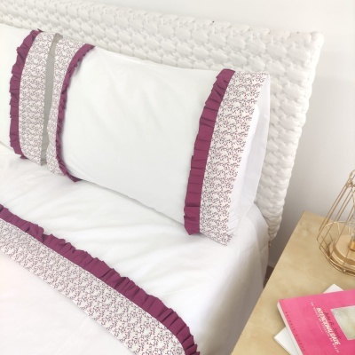 Capa almofada branca com folhos rosa e padrão floral rosa numa cama branca com cabeceira estofada e mesa de cabeceira com livros e candeeiro.