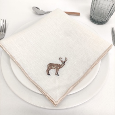 Guardanapo branco com bordado de veado sobre prato branco com talheres e copo