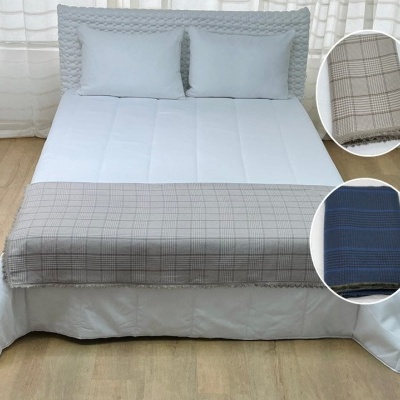 Cama com colcha branca e cobertor xadrez bege e azul no pé da cama em quarto com chão de madeira clara e cortinas brancas