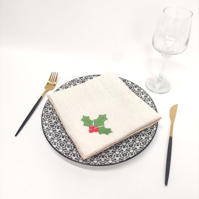 Mesa posta com prato cerâmico decorado, guardanapo de tecido com bordado de azevinho, talheres preto e dourado, e taça de vidro.