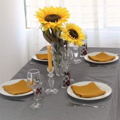 Mesa posta com toalha cinza e branca, pratos com guardanapos amarelos, copos com padrão floral, castiçal com vela amarela e vaso com girassóis