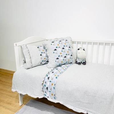 Cama branca infantil com roupa cinzenta e almofadas com padrão animal e peluche de vaca.