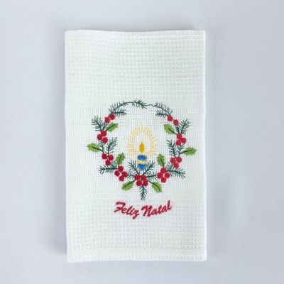 Toalha branca bordada com vela, coroa de folhas verdes e bagas vermelhas, e texto 'Feliz Natal' em vermelho