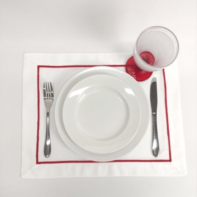 Jogo americano branco com borda vermelha, prato branco, talheres de metal e copo transparente com base vermelha