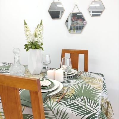 Mesa de madeira posta com toalha de folhas, louça branca, velas e vaso branco com flores, cadeiras de madeira e espelhos hexagonais na parede