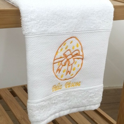 Toalha branca com bordado de ovo de Páscoa e texto pendurada em suporte de madeira.