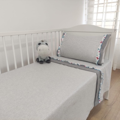 Cama individual branca com roupa de cama cinza e bordas florais, peluche de hipopótamo cinza ao lado, quarto com chão de madeira e cortina branca.