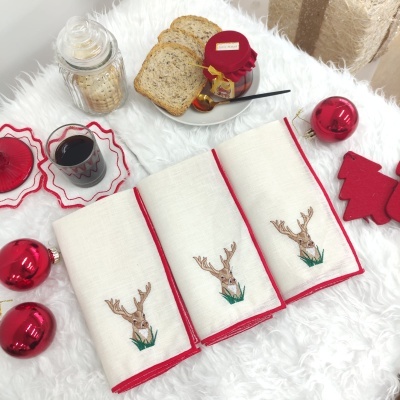 Três panos de cozinha brancos com bordas vermelhas e bordado de veado, pão, bolachas, enfeites de Natal em superfície branca felpuda.