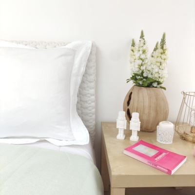 Cama com cabeceira branca, almofada branca e mesa de cabeceira com vaso de flores, figuras decorativas, vela e livro cor-de-rosa