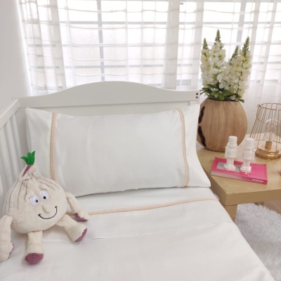 Cama branca com lençóis e fronhas bordadas bege, peluche em forma de alho, vaso de madeira com flores e velas ao lado.