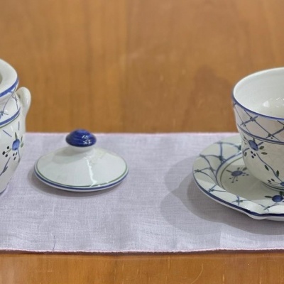 Conjunto de chávena, pires, recipiente com tampa e colher em porcelana branca e azul sobre toalha branca numa mesa de madeira.
