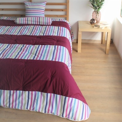 Colcha e almofada riscada colorida com vinho sobre cama de madeira clara e mesa de canto com vaso.