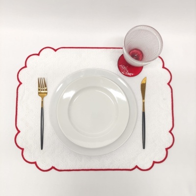Jogo de jantar com pratos brancos, talheres pretos e dourados, copo com base vermelha sobre individual branco com borda vermelha