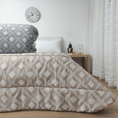 Cama com edredão e almofada em padrão de losangos cinza e bege, relógio de parede e mesa de cabeceira em quarto iluminado