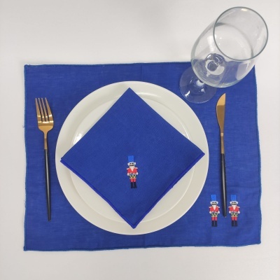 Conjunto de mesa azul com guardanapo e esteira bordados, prato branco, talheres dourados e copo transparente.