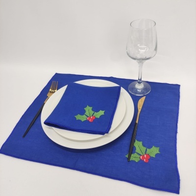 Lugar de mesa com guardanapo e marcador azul com bordado de folha de azevinho, prato branco, talheres dourados e copo de vidro transparente