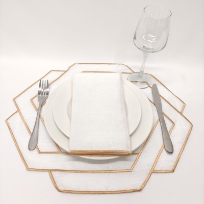 Mesa posta com jogo americano branco com borda dourada, prato branco, guardanapo, talheres e copo de vinho.