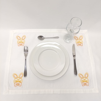 Jogo americano branco com bordado de coelhos amarelos, prato branco, talheres metálicos e copo de vidro