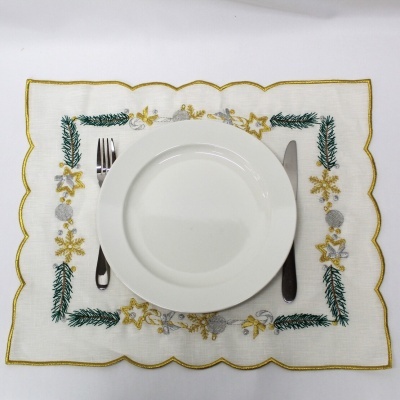 Jogo americano bordado em tecido branco com desenhos verdes, amarelos e prata, prato branco, garfo e faca.