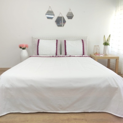 Cama de casal com colcha e fronhas brancas com detalhes roxos, cabeceira de madeira, três espelhos hexagonais na parede e vasos com flores