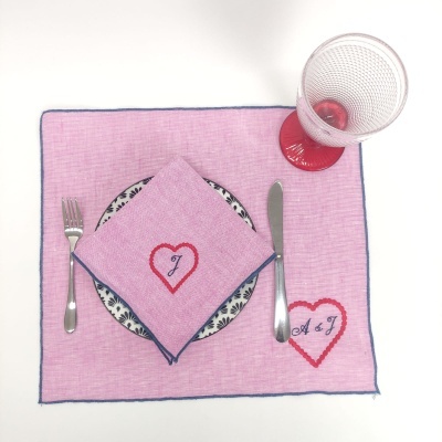 Conjunto de mesa com individual e guardanapo rosa, prato com padrão azul, talheres em aço inoxidável e copo transparente com pé vermelho.