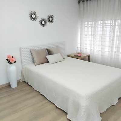 Quarto com cama branca e almofadas castanhas, vaso com flores e cortinas brancas