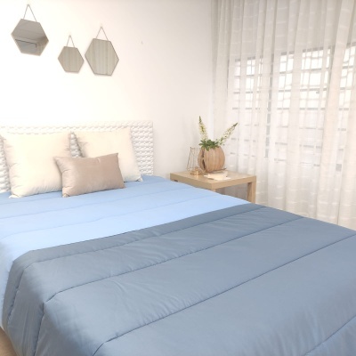Cama com edredão azul e almofadas, mesa de cabeceira com decoração e cortinas brancas