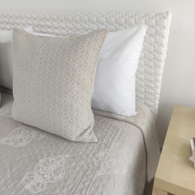 Cama com cabeceira branca acolchoada, almofadas bege e brancas, e roupa de cama cinzenta bordada com folhas brancas.