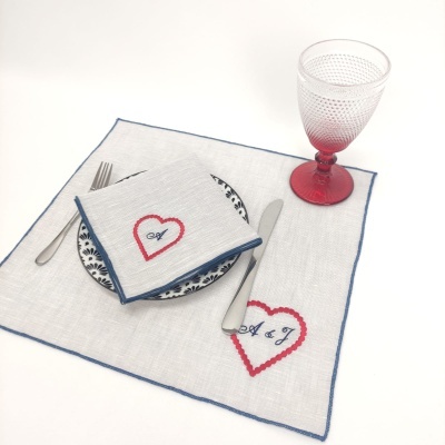 Jogo americano e guardanapo branco com bordado de coração vermelho e iniciais A & J, prato azul, talheres e copo vermelho.