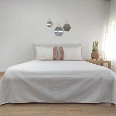 Cama de casal com cabeceira em rattan branco, colcha branca, almofadas decorativas e mesa de cabeceira com vaso e candeeiro